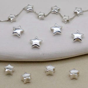 May include: Silver star-shaped beads scattered on a white surface, with some strung on a delicate chain. The beads have a shiny, reflective surface, and are evenly spaced. The image showcases jewelry components.
