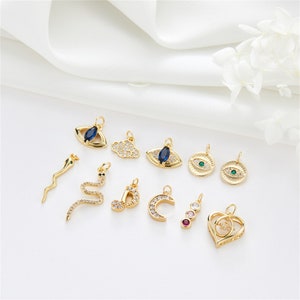 May include: Assortment of gold-tone charm pendants featuring various designs, including a snake, a cloud, a crescent moon, a musical note, a heart, and several evil eye designs with blue and green gemstones.