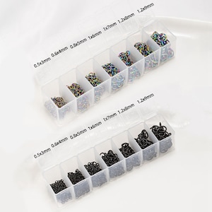 Op de afbeelding: Twee doorzichtige plastic organizers gevuld met kleine metalen verbindingsringen. Eén organizer bevat kleurrijke ringen, de andere zwarte. Maten zijn gelabeld: 0,5x3mm, 0,6x4mm, 0,8x5mm, 1x6mm, 1x7mm, 1,2x8mm en 1,2x9mm.