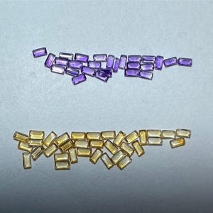 Puede incluir: Una colección de piedras preciosas rectangulares de color púrpura y amarillo. Las piedras preciosas están esparcidas sobre una superficie blanca.