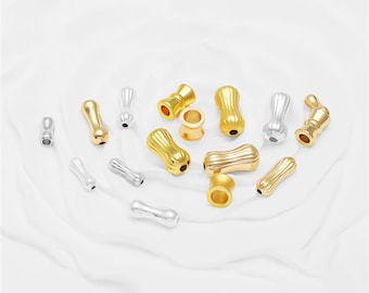May include: A collection of small, elongated, and hourglass-shaped beads in gold and silver tones. These metallic beads feature a ribbed, textured surface and a central hole, with some positioned vertically and others lying horizontally on the surface.