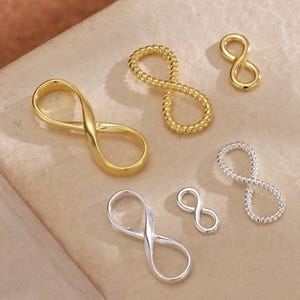 Könnte beinhalten: Eine Auswahl an Unendlichkeitszeichen-Anhängern in Gold- und Silberfarben. Die Anhänger haben unterschiedliche Designs, darunter glatte, gedrehte und kleine Größen. Die Anhänger werden auf einer neutralen, strukturierten Oberfläche gezeigt.