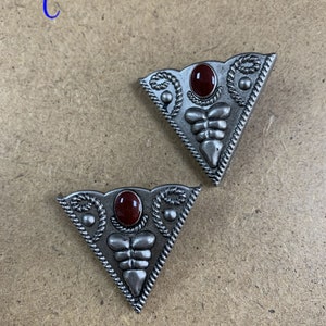 May include: Two silver-toned triangular metal buckles with a red stone in the center. The buckles have a decorative design around the stone and along the edges.