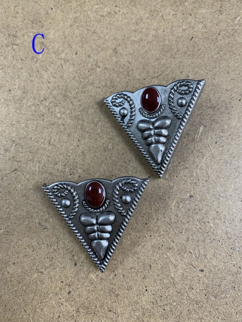 May include: Two silver-toned triangular metal buckles with a red stone in the center. The buckles have a decorative design around the stone and along the edges.