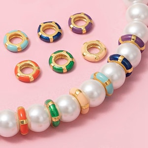 Puede incluir: Una colección de cuentas de esmalte y tono dorado de colores. Las cuentas son de varios colores, incluyendo azul, morado, verde, naranja y melocotón. Las cuentas son redondas con un centro dorado y se intercalan con grandes cuentas de perlas blancas.