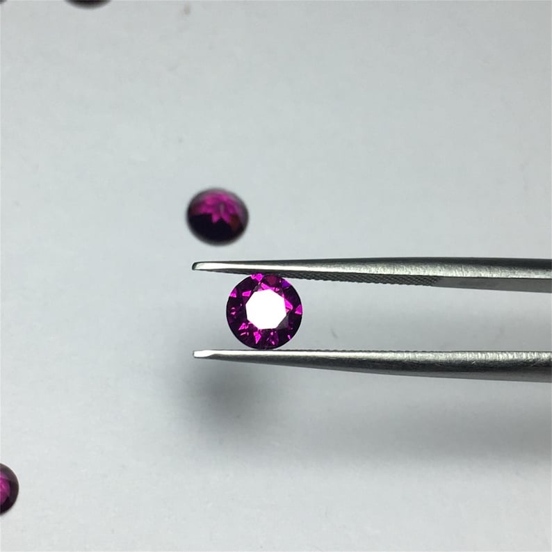 May include: A round, faceted, purple gemstone held by tweezers against a white background.