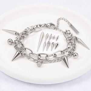 Könnte beinhalten: Ein silberfarbenes Punkrock-Armband mit mehreren Kettengliedern, konischen Spitzen und kleinen Kugelakzenten. Zusätzliche silberne Spitzen in verschiedenen Größen werden neben dem Armband angezeigt. Das Armband liegt auf einem weißen Teller.