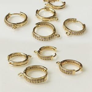 May include: Gold-tone hoop earrings, each adorned with tiny, clear stones. The earrings are circular with a hinged clasp. Multiple earrings are shown, highlighting their design and features.