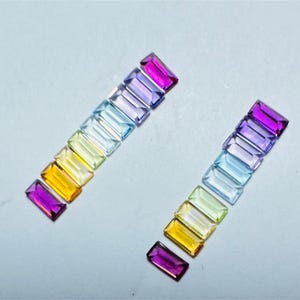 Puede incluir: Dos filas paralelas de piedras preciosas rectangulares con corte baguette en un espectro de colores que incluye magenta, morado, azul claro, amarillo pálido y naranja. Cada piedra presenta superficies facetadas que reflejan la luz.