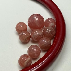 Pode incluir: Uma coleção de contas de pedras preciosas rosa e brancas, de tamanhos variados, está espalhada sobre uma superfície branca. Um objeto vermelho curvo emoldura as contas. As contas têm um acabamento polido e brilhante.