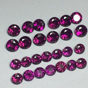 May include: A collection of round, faceted, purple gemstones. The gemstones are arranged in rows on a white surface.