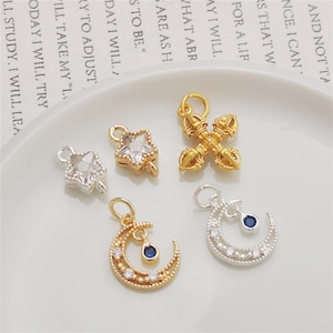 May include: A collection of five metal jewelry charms including two star-shaped charms with clear stones, one gold-toned vajra charm, and two crescent moon charms featuring small clear stones and a single dangling blue teardrop-shaped stone.