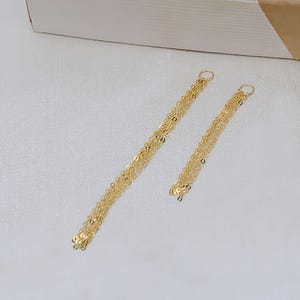 May include: A pair of gold-coloured chain earrings, each featuring multiple strands of delicate, flat, circular links. The earrings are presented on a white, textured surface, with a section of a white and gold box in the background.