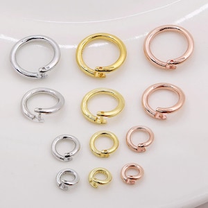 Puede incluir: Colección de anillos de resorte metálicos de varios tamaños en plata, oro y oro rosa. Los anillos están abiertos y tienen un pequeño mecanismo de cierre. Los anillos están dispuestos sobre una superficie blanca.