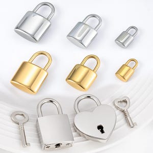 May include: An assortment of padlocks in various sizes and finishes. The collection includes silver and gold-colored padlocks, with one heart-shaped padlock and two keys. The padlocks are displayed on a white surface.