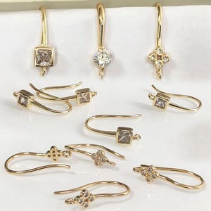 Pode incluir: Uma coleção de brincos dourados com vários designs. Alguns brincos apresentam pedras claras de corte quadrado, enquanto outros têm pedras claras redondas ou em forma de flor. Os brincos são exibidos em uma superfície branca.