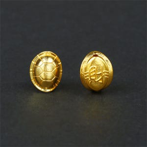 May include: A pair of gold-colored turtle-shaped earrings. One earring features a detailed turtle shell design, while the other has a Chinese character engraved on it.