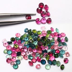 Può includere: Una collezione di gemme sfaccettate in vari colori, tra cui rosa, verde, blu e rosso. Un paio di pinzette tiene una gemma rossa. Le gemme sono sparse su una superficie bianca, mostrando la loro brillantezza.