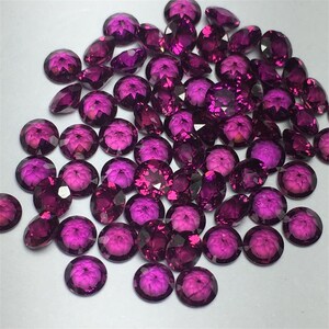 May include: A large group of round, faceted, purple gemstones. The gemstones are scattered on a white surface.