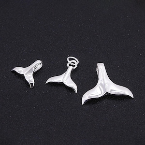 Può includere: Tre ciondoli a forma di coda di balena color argento di varie dimensioni. Ogni ciondolo ha una superficie lucida e riflettente, con un design curvo e simmetrico. I ciondoli sono esposti su uno sfondo scuro, evidenziando la loro forma e lucentezza.