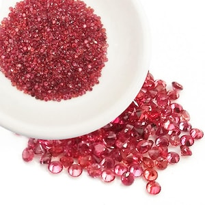 Puede incluir: Un cuenco de cerámica blanco rebosante de pequeñas gemas facetadas de color rojo intenso. Algunas gemas se han derramado del cuenco, esparcidas sobre una superficie blanca. Las gemas tienen un aspecto brillante y brillante.