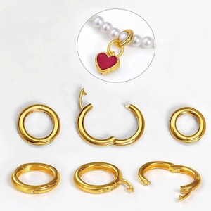 May include: Gold-colored jewelry clasps in various open and closed positions. A gold heart-shaped charm with a red enamel center is attached to a pearl necklace. The clasps are round and have a hinged design for easy opening and closing.