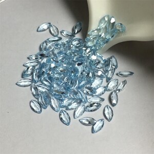 May include: A pile of light blue marquise-shaped gemstones, scattered on a white surface. The gemstones are faceted and have a brilliant shine.