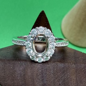 May include: A silver ring with an oval halo setting, surrounded by small, round, clear stones. The band is also set with smaller stones. The ring is displayed on a brown surface against a green background.