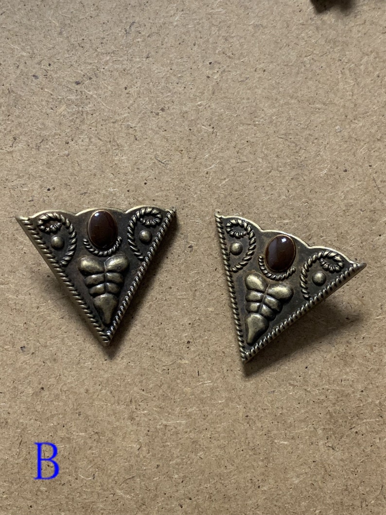 May include: Two gold-tone triangular metal embellishments with a brown stone in the center. Each embellishment has a decorative design around the stone and along the edges.