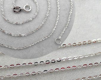 May include: Close-up of several delicate silver chain necklaces. The chains have a fine, oval link design and a shiny finish. One chain is coiled, and another has a small ring clasp. The chains are displayed on a light gray surface.