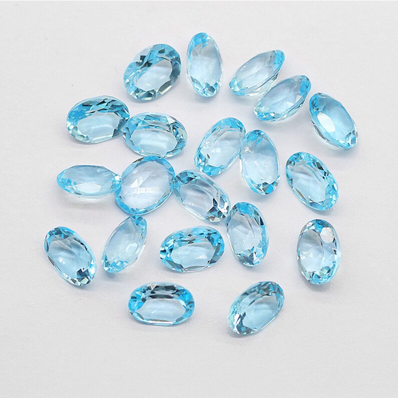 May include: A collection of oval-shaped, light blue gemstones. The gemstones are faceted and have a translucent appearance.