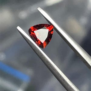 May include: A triangular red gemstone with facets, held by tweezers against a blurred background.