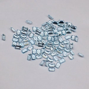 Puede incluir: Un montón suelto de piedras preciosas rectangulares facetadas de color azul claro. Las piedras preciosas están esparcidas sobre una superficie blanca.