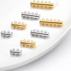 May include: An assortment of gold and silver-colored multi-strand jewelry clasps. The clasps have multiple loops for attaching strands and are displayed on a white surface. The clasps are cylindrical with rounded ends.