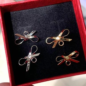 Pode incluir: Quatro pingentes de laço dourados em uma caixa de veludo vermelho. Os laços são de tamanhos diferentes e têm um laço na parte superior para prender a um colar ou pulseira.