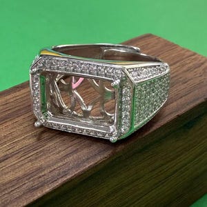 May include: A silver ring with a large rectangular setting encrusted with small clear stones. The ring is set on a wooden surface.