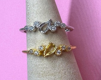 May include: Two rings, one silver and one gold, are shown. Both have a teardrop-shaped centre and small, round, clear stones. The bands have a textured, ridged design. The rings are set against a pink background.