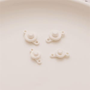 May include: Four small, off-white plastic fasteners, each featuring a central circular button mechanism with a raised dome and two flat, perforated tabs extending from opposite sides for attachment purposes, arranged in a two-by-two grid.