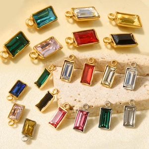 May include: A collection of rectangular charms featuring faceted crystals in various colors, including red, blue, green, yellow, pink, and clear, set in gold-toned or silver-toned metal frames with a small circular loop at the top for attachment.