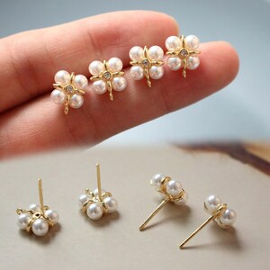 May include: Gold-tone stud earrings featuring a four-leaf clover design. Each clover is composed of four white pearls and a central clear crystal. The earrings have a post-back closure and are displayed on a light background.
