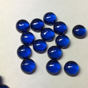 May include: A scattering of glossy, deep blue cabochon gemstones. The round, polished stones are displayed on a light surface. These gems are likely intended for use in jewellery or other decorative crafts.