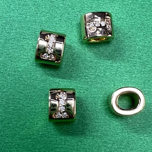 Pode incluir: Quatro contas de metal dourado com strass transparentes. Uma conta é uma simples forma de anel. As outras três contas são retangulares com uma fila de strass.