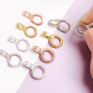 May include: Various small jewellery findings in gold, rose gold, and silver. Each finding has a circular design with a textured, rope-like pattern and a small, embellished bail. The findings are displayed on a light purple surface.