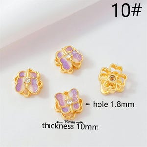 Könnte beinhalten: Vier goldfarbene, blütenförmige Charms mit einem lila-weißen Glitzerdesign. Jeder Charm hat einen kleinen, klaren Edelstein in der Mitte. Der Text "10#" befindet sich oben rechts. Die Dicke beträgt 10 mm, und das Loch ist 1,8 mm.