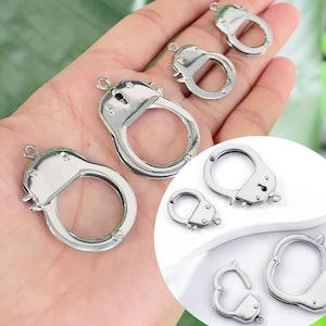 May include: Several silver-colored handcuff charms in various sizes. The charms have a detailed design, including the locking mechanism and a small loop for attaching to jewelry or other crafts. The handcuffs are displayed against a soft background.