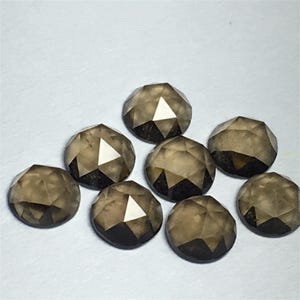 May include: Eight faceted, round-cut smoky quartz gemstones. The gems are a translucent brown color with a geometric pattern of triangular facets. The gemstones are arranged on a white surface.