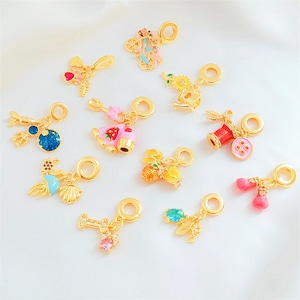 Può includere: Una collezione di ciondoli color oro con dettagli in smalto colorato. I ciondoli presentano vari disegni, tra cui una bicicletta, un cupcake, una bobina di cucito e una conchiglia. Ogni ciondolo ha un anello per il fissaggio a un braccialetto o una collana.