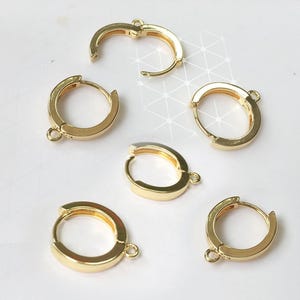 以下が含まれることがあります： ヒンジ開閉と小さなループが付いたゴールドカラーのフープピアス。ピアスは、さまざまな開閉位置で表示され、光沢のあるメタリック仕上げです。ジュエリーは白い表面に表示されています。