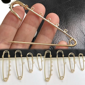 May include: A selection of gold-coloured safety pins and a large gold brooch. The brooch is held in a hand, highlighting its design. The safety pins vary in size and have small loops for attaching charms or beads. The items are displayed against a dark background.