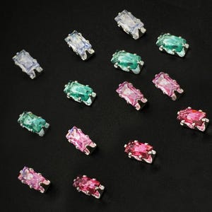 Puede incluir: Una colección de piedras preciosas de corte rectangular facetado engastadas en garras de metal plateado. Las piedras presentan varios colores translúcidos, incluyendo transparente, verde esmeralda y tonos rosados, sobre un fondo negro.